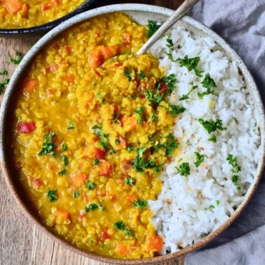 Bowl of creamy red lentil dahl with spices and herbs on a wooden table.